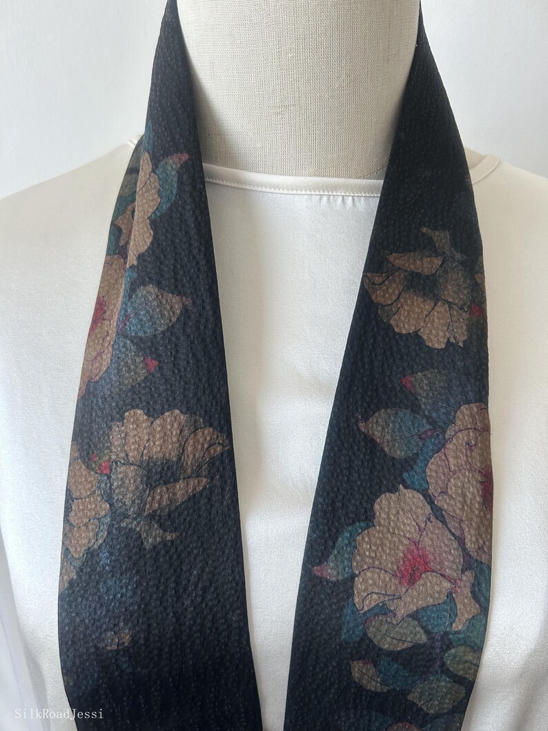 May include: A black silk scarf with a floral pattern in shades of brown, pink, and blue. The scarf is folded in half and draped over a white mannequin.