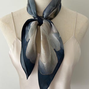 May include: A square scarf tied around the neck, featuring a watercolor-style floral pattern in shades of slate blue, charcoal gray, and muted beige. The scarf has a dark navy blue border and is displayed on a cream-colored camisole top.