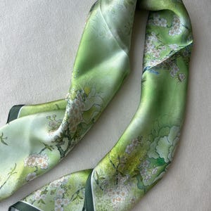 May include: A green silk scarf with a floral print of white blossoms and green leaves. The scarf has a dark green border.