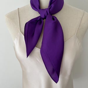May include: A vibrant purple square scarf tied in a simple knot around the neck of a mannequin. The scarf features a smooth, satin-like texture with a subtle sheen. The mannequin is wearing a plain, light cream-colored camisole with thin straps.