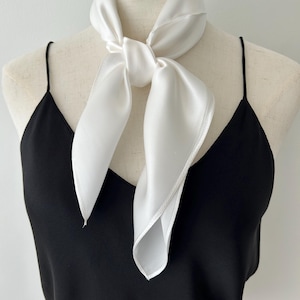 May include: A white satin scarf tied around the neck of a mannequin. The scarf is knotted in the front, with the ends hanging down. The mannequin is wearing a black spaghetti strap top.
