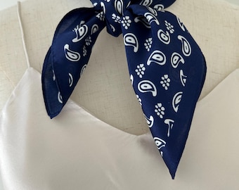May include: A dark navy blue square scarf featuring a repeating white paisley and floral pattern, tied in a knot around the neck. The scarf is displayed on a mannequin wearing a simple, light-colored, thin-strapped camisole top.