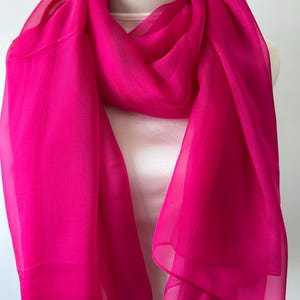 May include: A vibrant fuchsia scarf draped over a mannequin. The scarf is made of a sheer, lightweight fabric, with soft folds and a flowing design. The bright pink color is eye-catching and adds a pop of color.