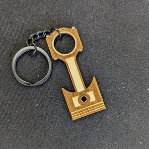 Puede incluir: Llavero de madera con forma de pistón de motor. El llavero es de madera marrón clara y oscura, con un llavero y una cadena plateados. El diseño del pistón incluye un agujero circular y líneas detalladas.