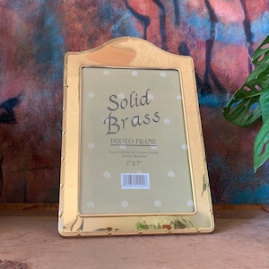 May include: A gold-colored, solid brass photo frame with a decorative design. The frame is 5 inches by 7 inches and has a white background with a floral pattern. The text "Solid Brass Photo Frame" is printed on the frame.