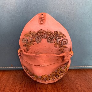 May include: A pink ceramic wall pocket with a decorative floral design and a small face sculpted on the top. The pocket has a scalloped edge and a small opening at the top.