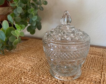 May include: A clear glass jar with a lid, featuring a diamond pattern and a knob on top. The jar is sitting on a woven mat.