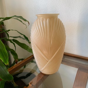 May include: A pale peach glass vase with a fan-shaped design. The vase is sitting on a wooden table with a glass top.