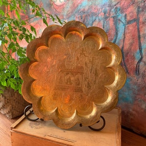 May include: A large, round, brass serving tray with a scalloped edge. The tray has an intricate, engraved design featuring a central motif of a temple or pavilion with figures. The tray is displayed on a wooden box with the text "Please handle carefully".