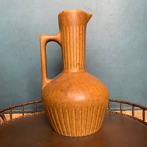 May include: A brown ceramic pitcher with a textured surface and vertical lines. The pitcher has a wide base and a narrow neck with a spout.