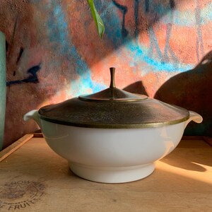 May include: A white ceramic serving bowl with a brass lid and two handles. The bowl is sitting on a wooden surface with the words "Please handle carefully" written on it.