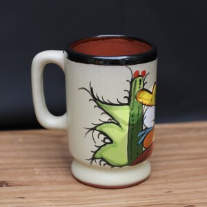 May include: A white ceramic mug with a brown rim and a black handle. The mug has a hand-painted design of a cactus and a person wearing a sombrero.