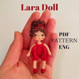May include: A handmade crochet doll named "Lara Doll" with brown curly hair and a red outfit. The doll has a red bow, rosy cheeks, and is held in a hand. The image also includes the text "PDF PATTERN ENG".