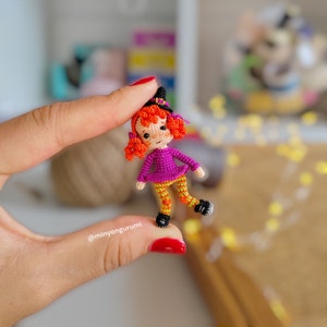 May include: A small crocheted doll wearing a purple dress, orange and yellow striped pants, and a black witch hat. The doll has orange hair and a friendly face.