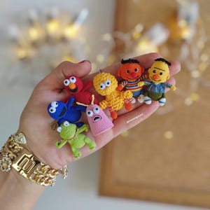 May include: A collection of small, crocheted Sesame Street character dolls in various colors. The characters include Elmo, Cookie Monster, Bert, Ernie, and others. The dolls are held in a hand, showcasing the detailed craftsmanship and vibrant colors.