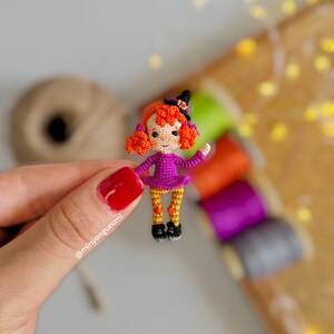 May include: A small crocheted witch doll with orange hair, a purple dress, orange and yellow striped leggings, and a black witch hat. The doll is holding a small broom.