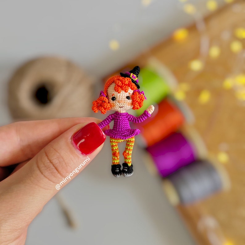 May include: A small crocheted witch doll with orange hair, a purple dress, orange and yellow striped leggings, and a black witch hat. The doll is holding a small broom.