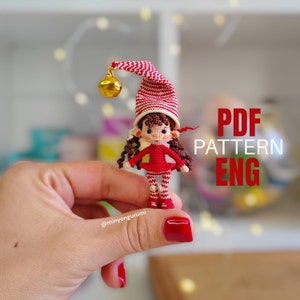 May include: A crocheted elf doll wearing a red and white striped hat with a gold bell. The doll is holding a red dress and has brown hair. The text "PDF PATTERN ENG" is visible in the background.