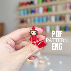 May include: A crocheted amigurumi elf doll wearing a red and white hat. The doll is holding a small green leaf in its hand. The image includes the text "PDF PATTERN ENG".