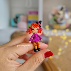 May include: A small crocheted witch doll with orange hair, a purple dress, and green and orange striped leggings. The doll is wearing a black witch hat and is holding a small broom.