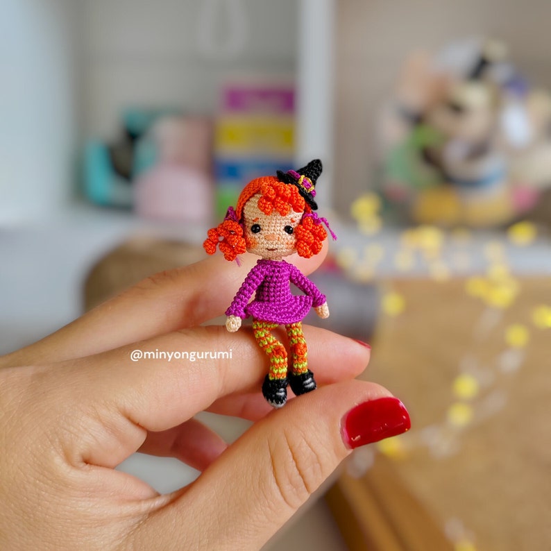 May include: A small crocheted witch doll with orange hair, a purple dress, and green and orange striped leggings. The doll is wearing a black witch hat and is holding a small broom.