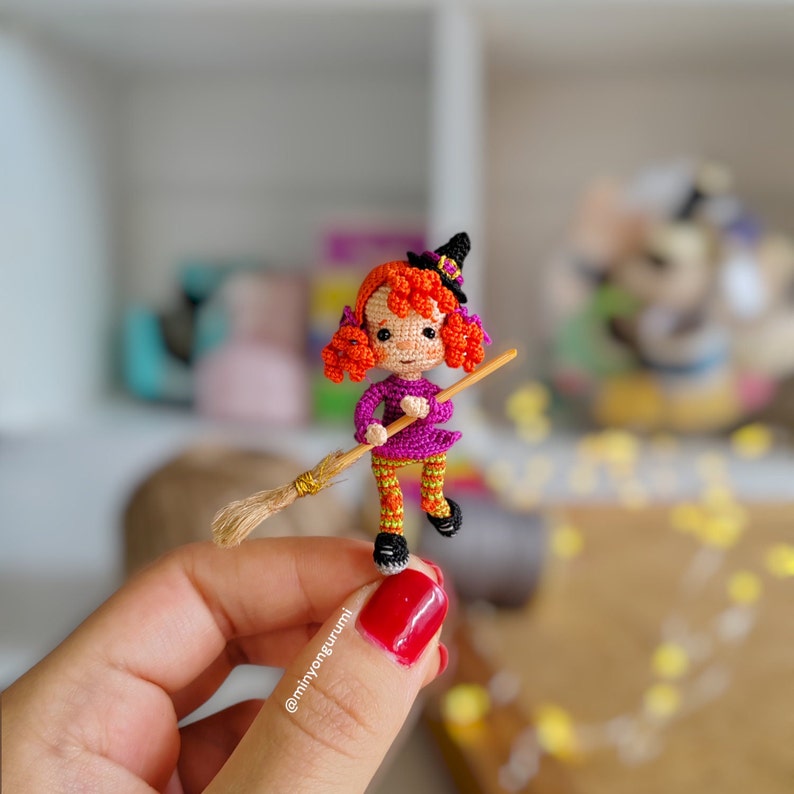 May include: A small crocheted witch doll with orange hair, a purple dress, and orange and yellow striped tights is holding a miniature broom. The doll is wearing a black witch hat with a gold buckle. The doll is being held by a hand with red nail polish.