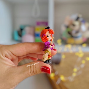 May include: A small crocheted witch doll with orange hair, a purple dress, and a brown broom. The doll is holding the broom in its right hand and is sitting on the broom. The doll is about 2 inches tall.