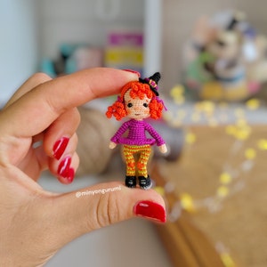 May include: A small crocheted witch doll with orange hair, a purple dress, and orange and yellow striped pants. The doll is wearing a black witch hat and is holding a small broom.