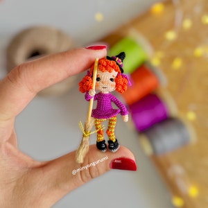May include: A small crocheted witch doll with orange hair, a purple dress, and orange and yellow striped tights. The doll is holding a small brown broom and wearing a black witch hat. The doll is being held in a hand.