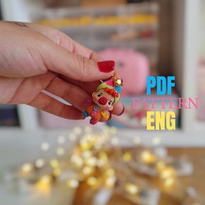 Puede incluir: Una pequeña muñeca de payaso de ganchillo colorida con una nariz roja, un sombrero verde y azul y un dije dorado. La muñeca se sostiene en una mano con esmalte de uñas rojo. El texto "PDF PATTERN ENG" es visible en el fondo.