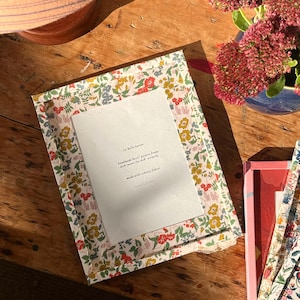 Puede incluir: Un marco de fotos rectangular cubierto con una tela floral con pequeñas flores amarillas, rojas y verdes sobre un fondo blanco. Un inserto de papel blanco está centrado en el marco con el texto La Belle Saison, handmade 8x10 picture frame with insert for 8x8 artwork, made with Liberty fabric.