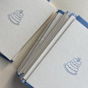 Op de afbeelding: Een stapel witte verjaardagskaarten met een blauwe taartillustratie en blauwe randen. De taart heeft drie kaarsen. De kaarten zijn schuin opgestapeld, met enkele kaarten uitgespreid.
