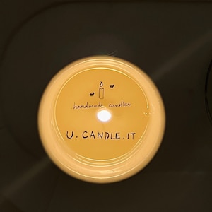 May include: A lit candle with a yellow wax surface. The candle has the text "U. CANDLE. IT" and "handmade candles" written on it. The candle has a small candle and heart illustration.