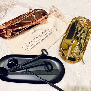 May include: A set of candle care tools in three different finishes: rose gold, black, and gold. The tools include a wick trimmer, candle snuffer, and wick dipper. The tools are arranged on three separate trays. The trays are oval shaped and made of metal. The trays are labeled "Candle Care" and "Wick Trimmer, Candle Snuffer, Wick Dipper".