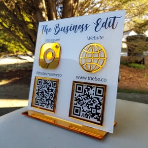 May include: White acrylic sign with gold accents featuring the text "The Business Edit". The sign includes Instagram and website information, along with QR codes for both.