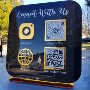 May include: Black sign with gold trim and the text "Connect With Us". The sign has four sections with icons and text. The top left section has an Instagram icon and the text "Instagram". The top right section has a QR code and the text "@krujuicebar". The bottom left section has a globe icon and the text "Website". The bottom right section has a QR code and the text "krujuicebar.com".