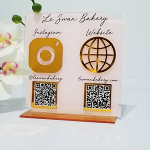 Puede incluir: Un letrero de acrílico rosa con letras doradas que dice "Le Swan Bakery". El letrero tiene dos secciones, una para Instagram y otra para el sitio web. Cada sección tiene un icono dorado, un código QR y texto. La sección de Instagram tiene un icono de Instagram dorado, el texto "@leswanbakery" y un código QR. La sección del sitio web tiene un icono de globo dorado, el texto "leswanbakery.com" y un código QR.
