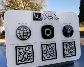 May include: White sign with black text and icons. The text reads "Violeta Giovanna" and includes the website address "https://www.violeta-giovanna.com", Instagram handle "@violeta_giovanna_official", and Facebook handle "@VioletaGiovannaOfficial".  Each social media platform has a corresponding icon and QR code.