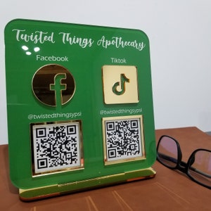 Może przedstawiać: Zielony znak akrylowy z pozłacanymi krawędziami, z napisem "Twisted Things Apothecary". Znak ma dwie sekcje, jedną z logo Facebooka i kodem QR, a drugą z logo TikToka i kodem QR. Znak stoi na drewnianej powierzchni.
