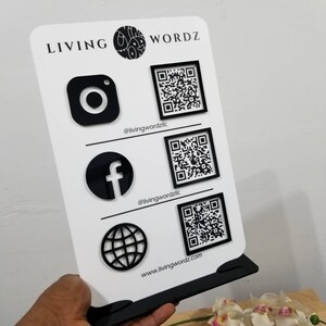 May include: A white sign with black text and graphics. The text reads "LIVING WORDZ". The sign has icons for Instagram, Facebook, and a globe, each with a corresponding QR code. The text below the icons reads "@livingwordzllc" and "www.livingwordz.com".