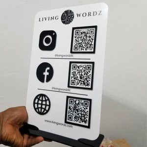 May include: White sign with black text that reads "LIVING WORDZ". The sign has three sections with black icons and QR codes. The icons are Instagram, Facebook, and a globe. The text below the icons reads "@livingwordzllc", "@livingwordzllc", and "www.livingwordz.com".