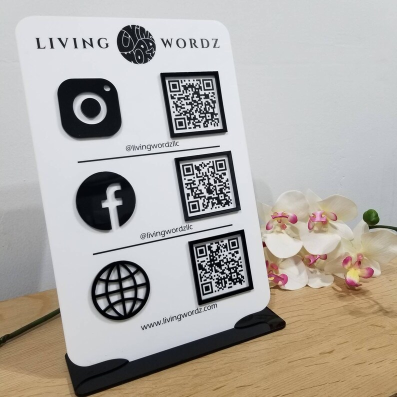 May include: White sign with black text and graphics. The text reads "LIVING WORDZ" and includes social media icons for Instagram, Facebook, and a website. Each icon has a corresponding QR code.