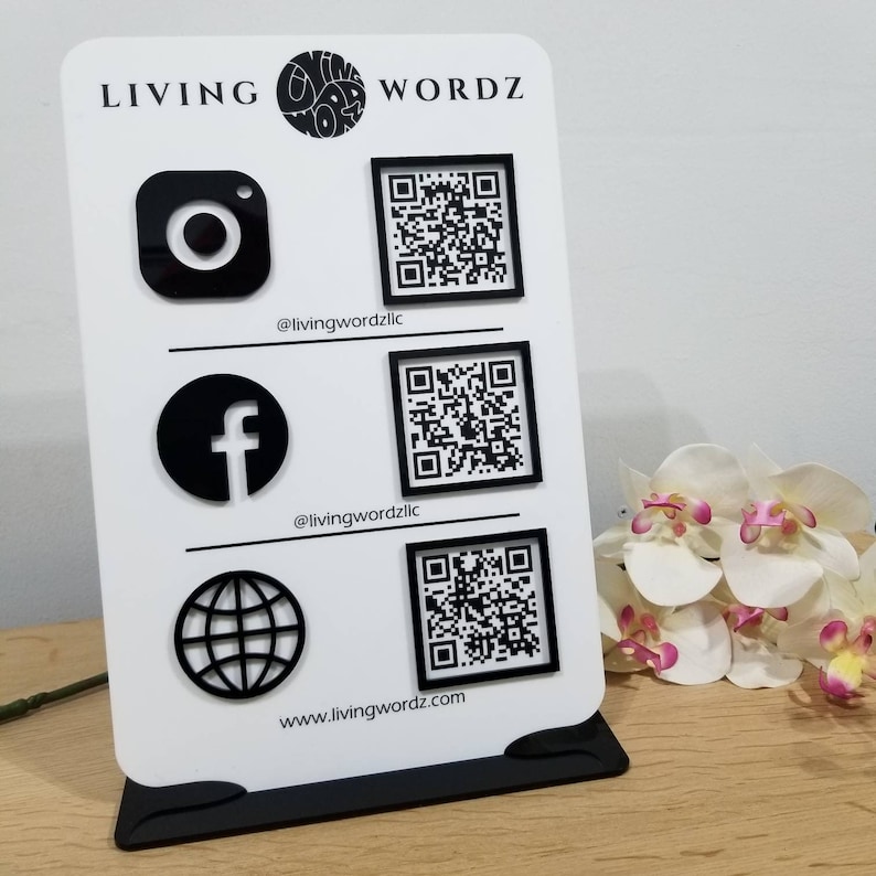May include: A white sign with black text and graphics. The text reads "LIVING WORDZ". The sign has three rows of icons and QR codes. The first row has an Instagram icon and a QR code. The second row has a Facebook icon and a QR code. The third row has a globe icon and a QR code. The text below the icons reads "@livingwordzllc" and "www.livingwordz.com".