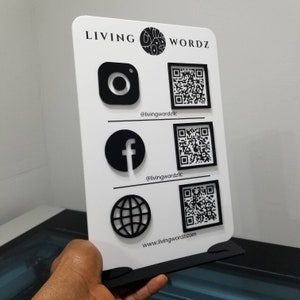 May include: White sign with black text and icons. The text reads "LIVING WORDZ" and includes social media handles and a website address: @livingwordzllc, @livingwordzllc, www.livingwordz.com. The icons include Instagram, Facebook, and a globe.