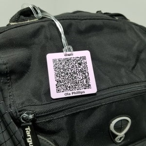 Könnte beinhalten: Ein rosa Gepäckanhänger mit einem schwarz-weißen QR-Code und dem aufgedruckten Text "Ola Phillips".