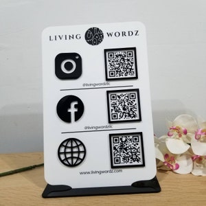 May include: White sign with black text and icons. The text reads "LIVING WORDZ". The icons are Instagram, Facebook, and a globe. Each icon has a black and white QR code next to it. The text below the icons reads "@livingwordzllc", "@livingwordzllc", and "www.livingwordz.com".