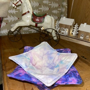 Peut inclure: Un protège-bol matelassé violet et blanc avec un motif tie-dye. Le protège-bol est plié et posé sur une surface en bois.