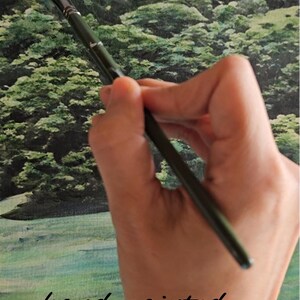 Op de afbeelding: Een hand houdt een zwart penseel boven een groen-blauw aquarel schilderij. Het schilderij toont een landschap met bomen en gras. De tekst "hand-painted" is in zwart onder de kwast geschreven.