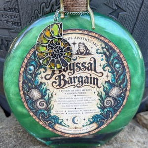 May include: A round green glass bottle with a cork stopper wrapped in twine and gold wax. A silver-toned ammonite pendant with iridescent green and gold inlay hangs from the neck. The label reads Abyssal Bargain, A Potion of Deep Secrets & Hidden Power.