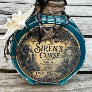 May include: A round glass bottle filled with teal liquid, featuring a label that reads Siren's Curse, Rare & Ocean-Haunted, Best Left Unsealed, Two Toads Apothecary. The neck is wrapped in twine and black wax, adorned with a white starfish charm.